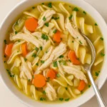 close-up of homemade chicken noodle soup with rotisserie chicken, egg noodles, carrots, celery, and parsley in a white bowl